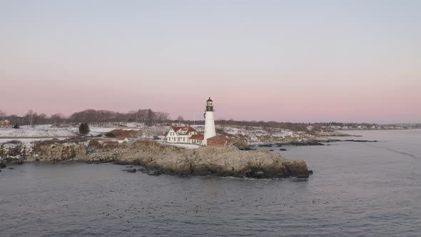 Lighthouse on rocky coastal outcrop above floating sea ducks, winter sunrise, SLOW AERIAL ORBIT