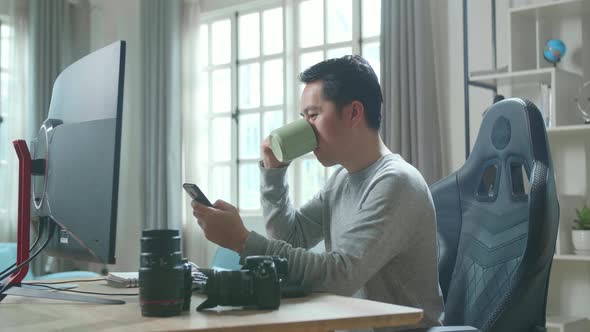 Asian Cameraman Using Mobile Phone And Drinking Coffee While Using Computer For Working At Home