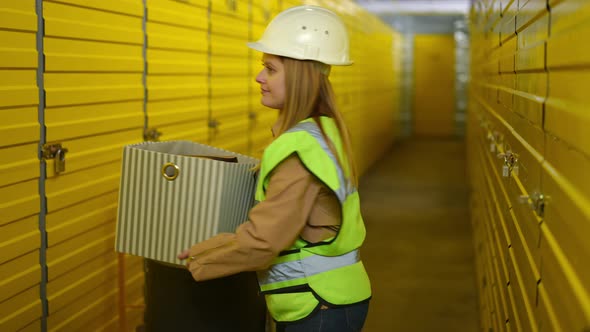 Serious Young Woman in Uniform and Hard Hat Walking Away with Box in Warehouse