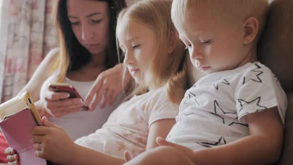 Young Mother, Daughter and Little Son Are Using Gadgets Lying on a White Bed. Concept Modern Family