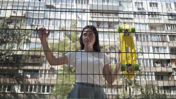 Portrait of Cute Woman with a Skateboard Looking at the Camera Standing Behind the Mesh Fence