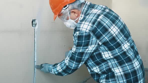 An Elderly Male Worker in a Helmet Mask and Goggles Breaks the Wall with a Chisel and Hammer to Wire