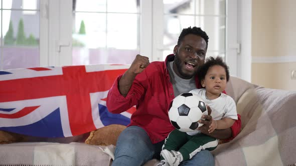 Front View African American Father and Toddler Son Watching Soccer Match Sitting at Home with