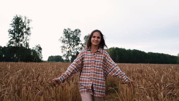 Happy Brunette Girl in a Shirt Walks Across the Field and Touches Spikelets with Her Hands