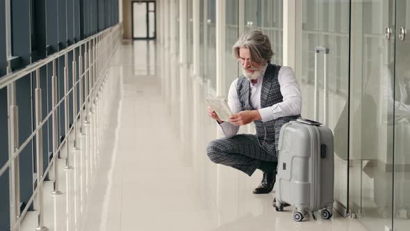 Senior Man Planning Vacation with a Map Sitting with a Suitcase at the Airport