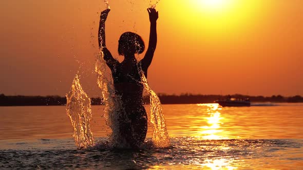 Silhouette of Woman at Sunset Raises Hands Up and Creating Splashes of Water. Slow Motion
