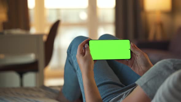 Woman at Home Lying on a Sofa and Using Smartphone with Green Mock-up Screen in Horizontal Mode