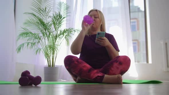 Fitness Training Blonde Overweight Woman Sitting on Yoga Mat and Looking at Her Phone While Drinking