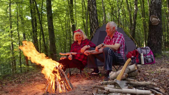 Senior Elderly Grandmother Grandfather Cooking Frying Sausages Over Campfire in Wood at Camping