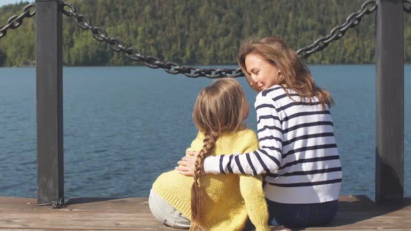 Loving Mother Hugs Her Daughter Sitting on a Pier Near a Beautiful Lake