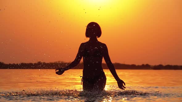Silhouette of Woman at Sunset Raises Hands Up and Creating Splashes of Water