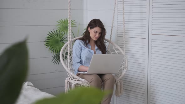 Girl Sitting with a Laptop on Her Lap in a Cozy Hanging Chair. Young Female Freelancer Working