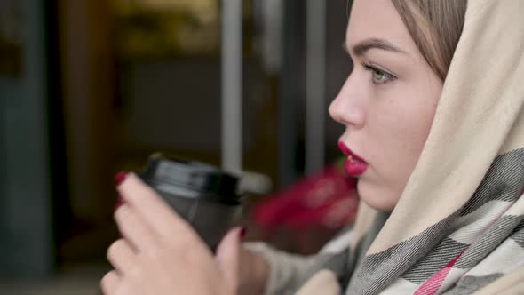 woman straightens her scarf on her head and drinks coffee from a disposable cup