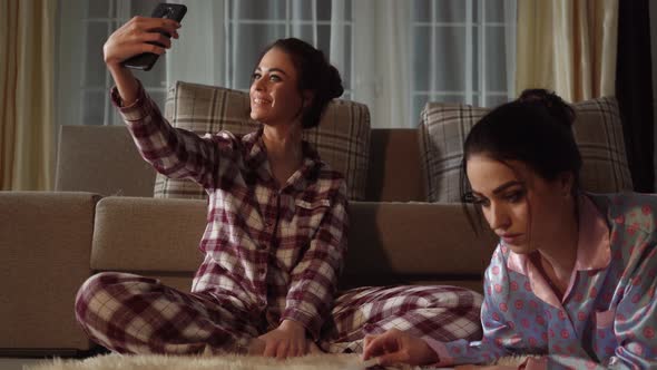 One of Two Pretty Sisters Making Selfie on Smartphone While Another Reading Magazine in Cozy Living Room