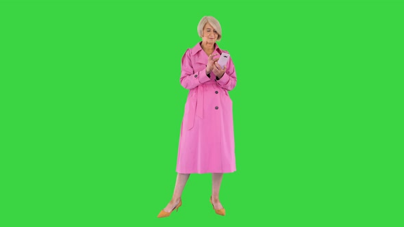 Cheerful Senior Lady in Pink Trench Coat Using Her Smartphone on a Green Screen Chroma Key