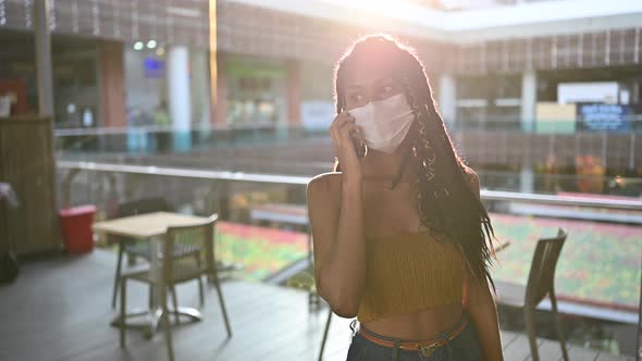Young black woman in face mask speaks on smart phone in commercial mall