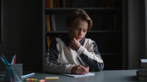 Portrait of Pupil Boy Thoughtful Holding Hand on Chin Writing in Notebook with Pen Sitting at Desk