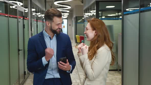 Excited Businessman and Businesswoman Rejoices at Message on Smartphone Standing at Office