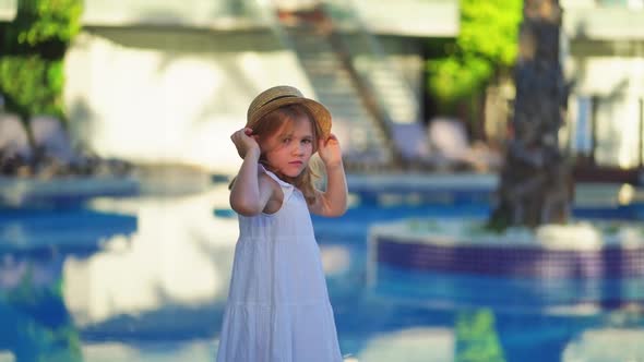 Cute Little Girl in a White Dress and Straw Hat By the Pool Near the Villa