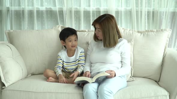 Asian Mother And Her Son Reading A Book On Sofa In Living Room