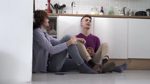 Gay Couple Sitting on the Kitchen Floor Having a Romantic Conversation While Drinking a Hot Drink