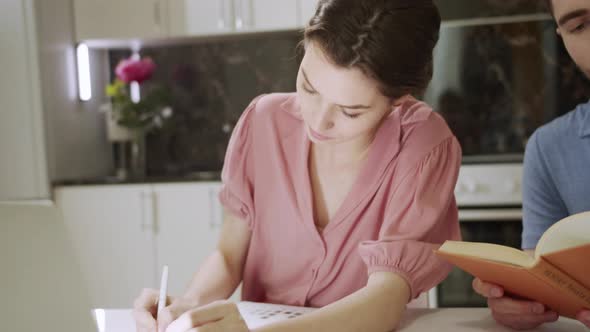 A Young Man is Reading a Book While His Wife is Writing Something Down on Paper