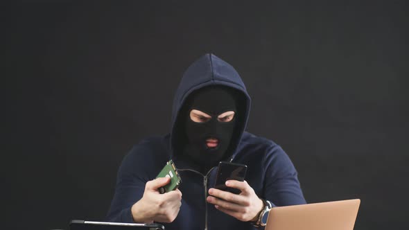 Male Hacker in Black Mask and Hood Uses Laptop To Hack a Hard Drive.