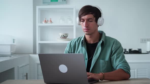 Young Man Freelancer in Headphones Starting Working at Laptop While Sitting at Kitchen Table at Home