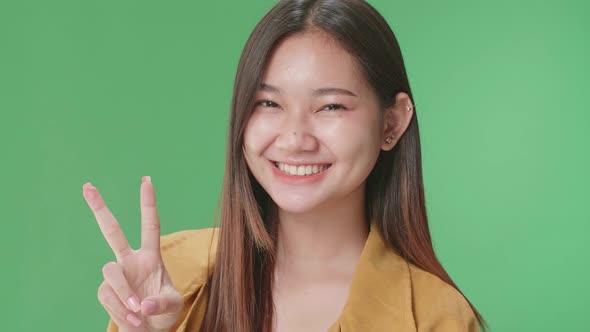 Smiling Young Asian Woman Showing Gesture Peace While Standing On Green Screen In The Studio