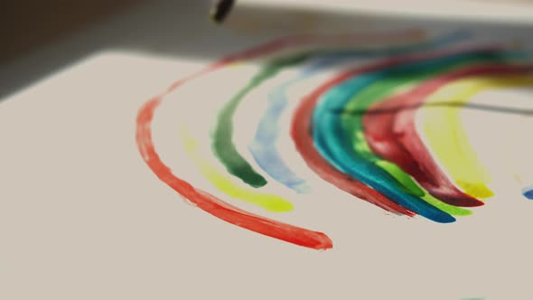 Little Girl Holds a Brush in Her Hand and Paints a Rainbow with Colored Paints on White Paper