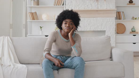 Bored Lonely Young African American Woman Sitting on Couch in Living Room Holding TV Remote Control