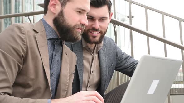 Two young bearded men sitting outdoor discussing using personal computer