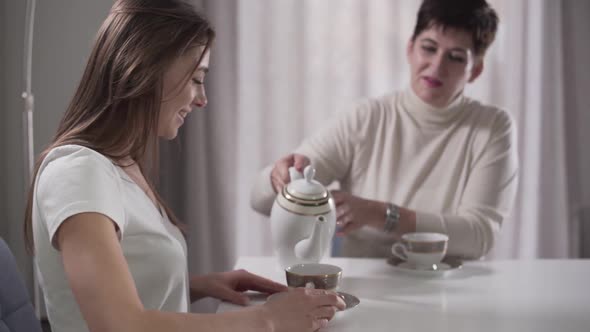 Side View of Young Caucasian Girl Sitting at the Table As Her Good-looking Mother Pouring Tea Into