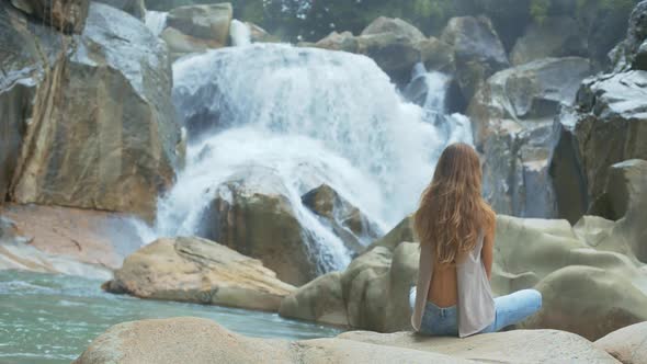 Girl Sits in Lotus Pose Against Waterfall and Rocks