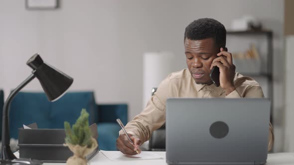 Adult Black Man is Communicating By Mobile Phone Sitting at Table with Laptop Writing Notes Working