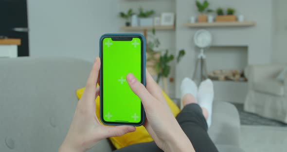 Woman at Home Lying on a Couch Using Smartphone with Green Mock-up Screen, Doing Swiping, Scrolling