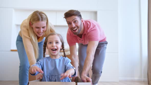 Joyful mother and father move brown cardboard box with funny little preschooler daughter