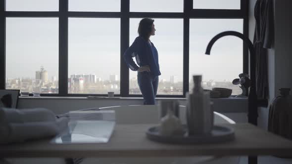 Long Shot of Brunette Caucasian Woman Looking Out the Window and Sitting Down on Windowsill