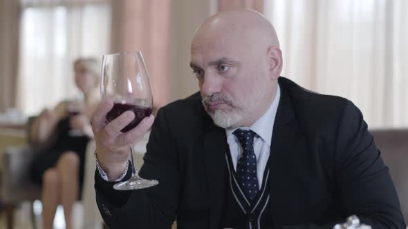 Close-up Portrait of Confident Bald Caucasian Man Drinking Red Wine in Restaurant As Blurred Blond