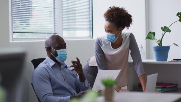 Diverse businessman and businesswoman in face masks discussing and using laptop in office