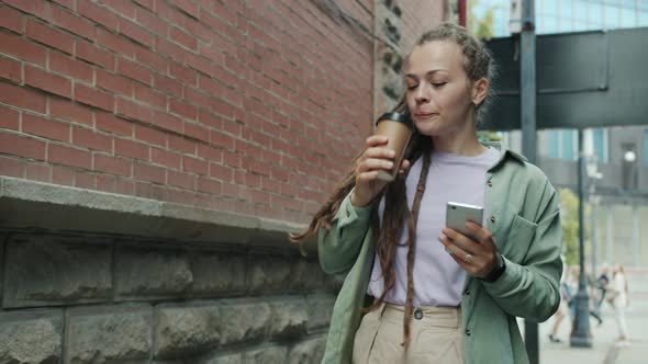 Beautiful Girl with Dreads Drinking to Go Coffee and Using Smart Phone Walking Outside Alone