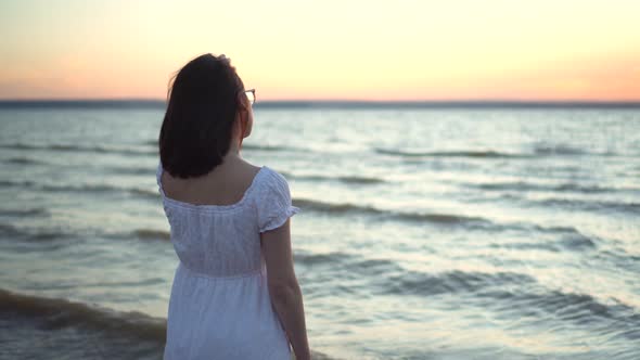 Attractive Young Woman Looks at the Sunset on the Beach By the Sea. The Girl in a White Dress Stands