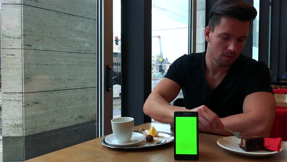 A Young Man Sits at a Table in a Cafe, Eats Pie and Drinks Tea, a Smartphone with a Green Screen