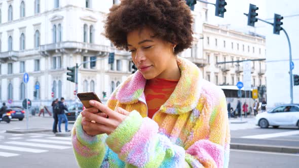 Young beautiful curly hair black woman outdoor in the city smiling using smartphone