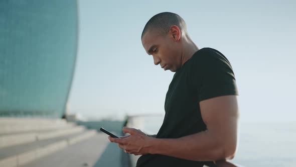Positive bald sportsman wearing black t-shirt texting on mobile outdoors