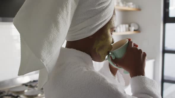 Happy african american woman with beauty mask on face, drinking coffee in kitchen