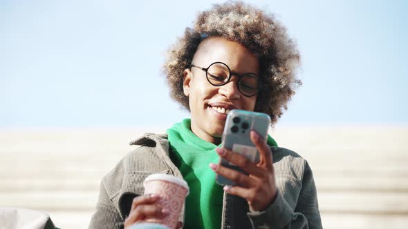 Cheerful adult African woman wearing casual clothes looking at phone and drinking coffee