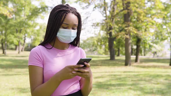 A Young Black Woman in a Face Mask Works on a Smartphone and Acts Frustrated in a Park