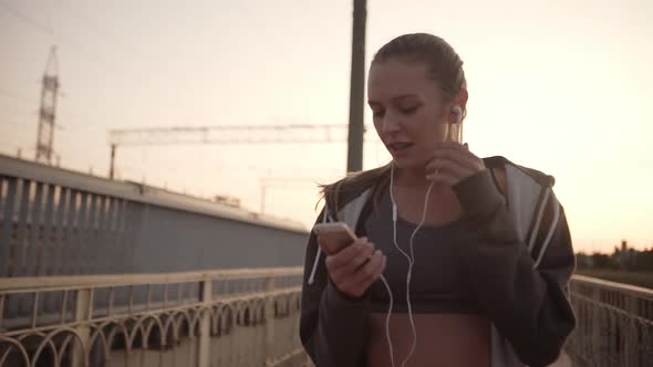 Smiling Girl with Tied Blond Hair Taking Break After Running Along City Bridge at Sunset with Moving