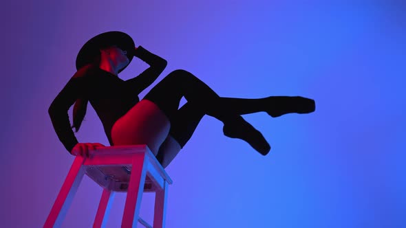 Professional Ballet Dancer in Hat Sitting on High Chair on Multicolored Background Under Violet Neon